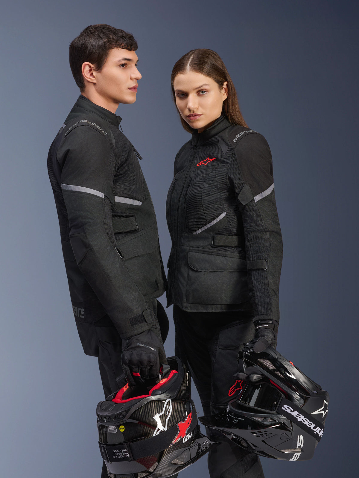 Male and female riders in Alpinestars motorcycle gear, featuring the Stella Andes Air Drystar® Jacket in Black, matching textile riding pants and gloves, both holding black and white helmets, studio shot showing coordinated adventure touring apparel