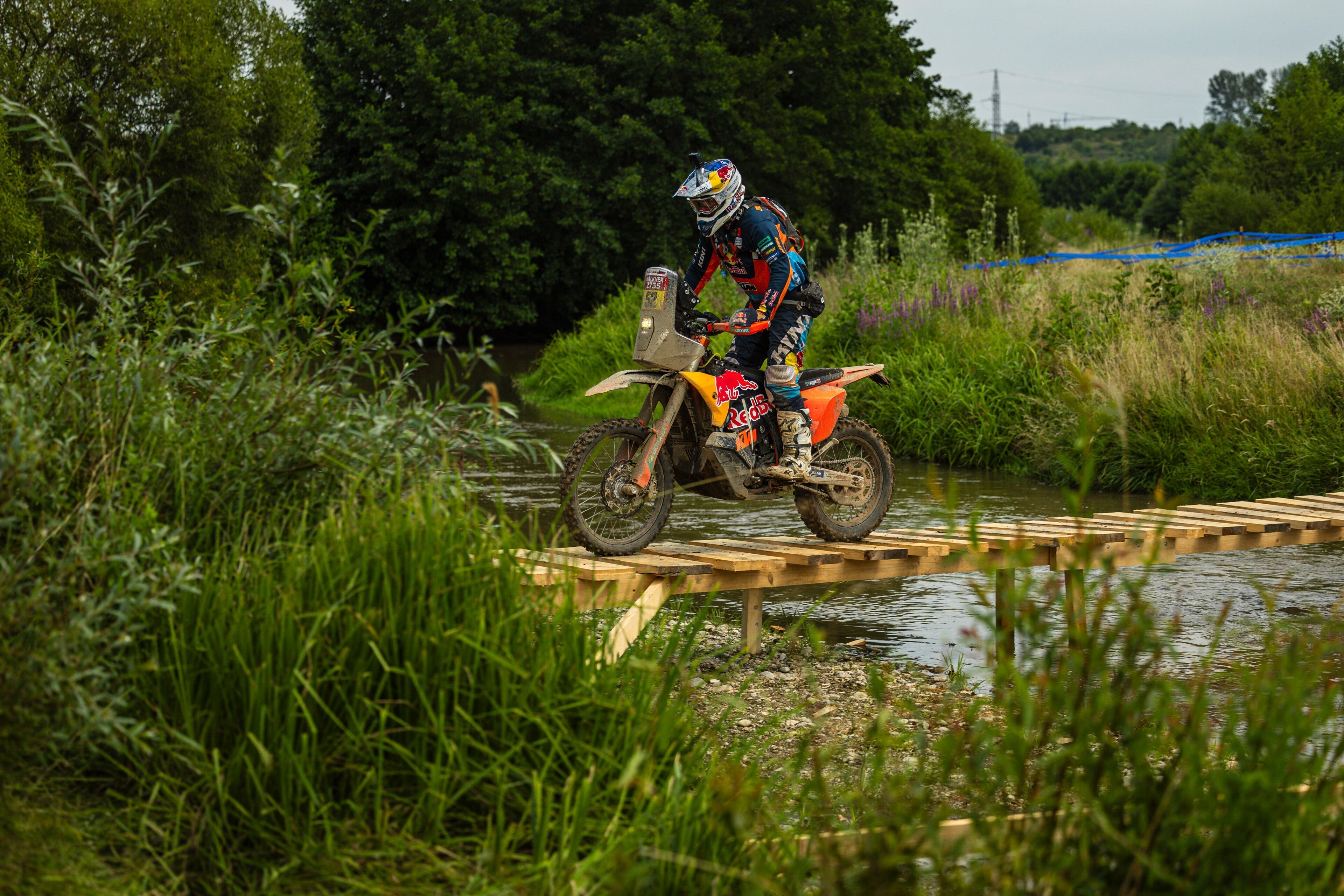Matthias Walkner riding a dirt bike on a wooden platform over water with greenery in the background