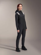 Woman wearing the Alpinestars Stella A-Dura Rain Jacket in Black, 2.5-layer waterproof MTB shell with light grey shoulder accents, matching black pants and cycling shoes, studio portrait for performance cycling gear