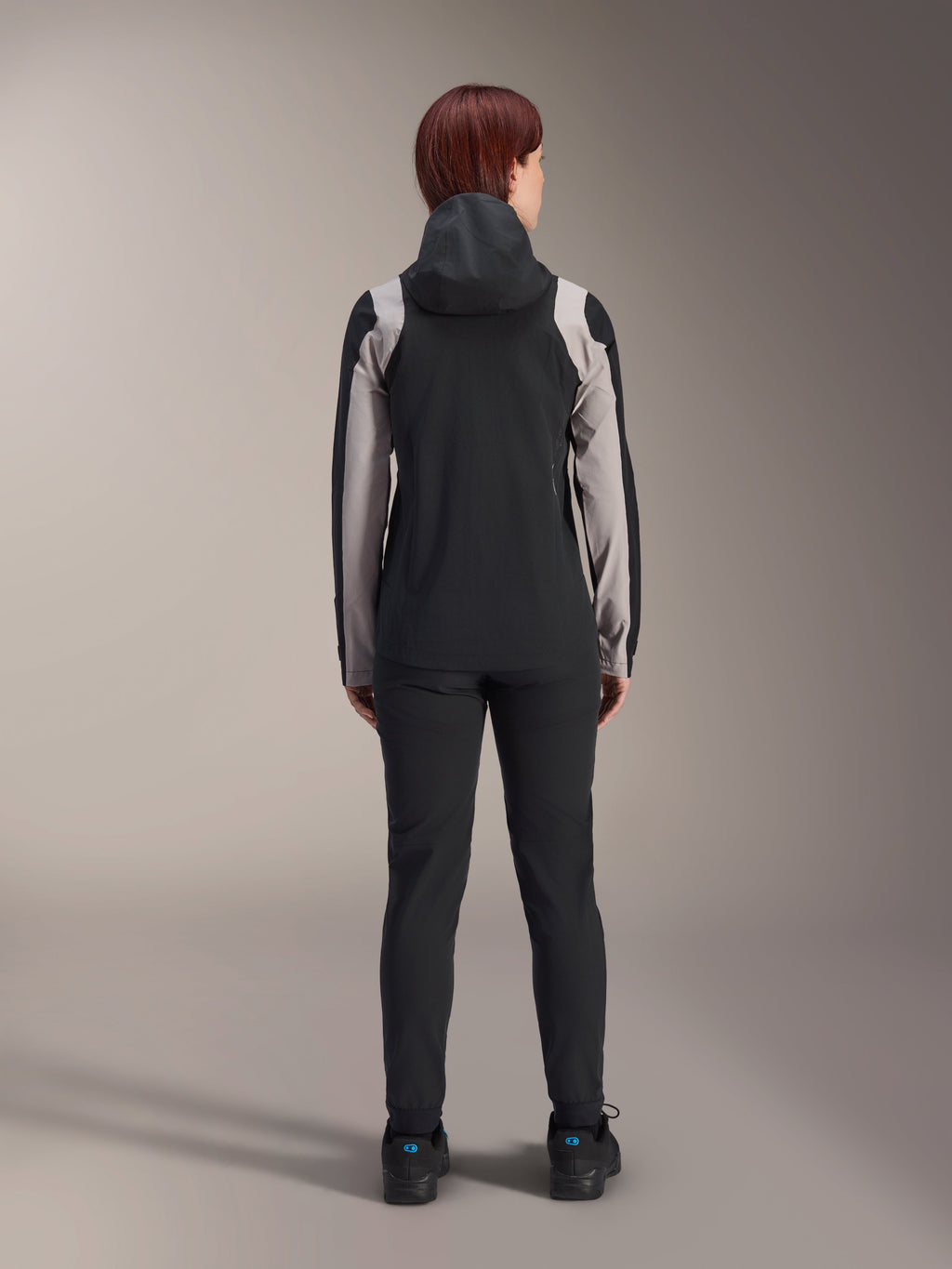 Female rider from behind wearing Alpinestars Stella A-Dura Rain Jacket, Black with light grey panels on the sleeves and shoulders, hood up, worn with black pants and cycling shoes, technical mountain bike rain gear