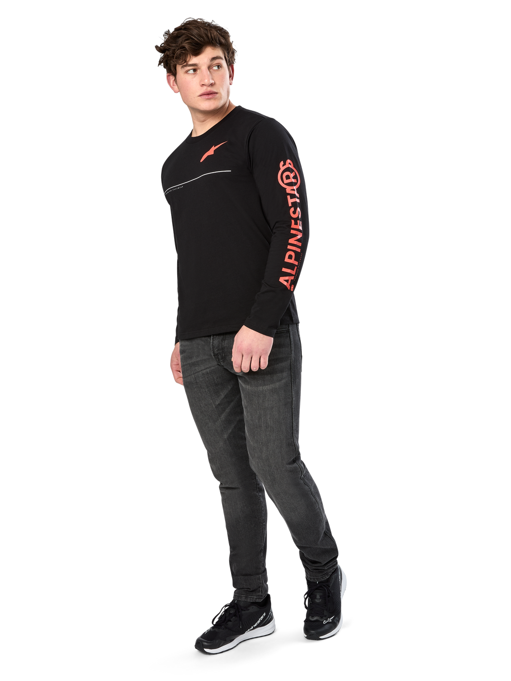 Male model wearing Alpinestars Committed CSF Tee, Long Sleeve T-Shirt, Black, orange-red chest logo and sleeve branding, paired with dark gray jeans and black sneakers, casual motorsport-inspired streetwear