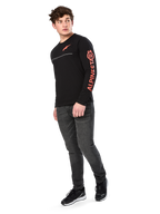 Male model wearing Alpinestars Committed CSF Tee, Long Sleeve T-Shirt, Black, orange-red chest logo and sleeve branding, paired with dark gray jeans and black sneakers, casual motorsport-inspired streetwear