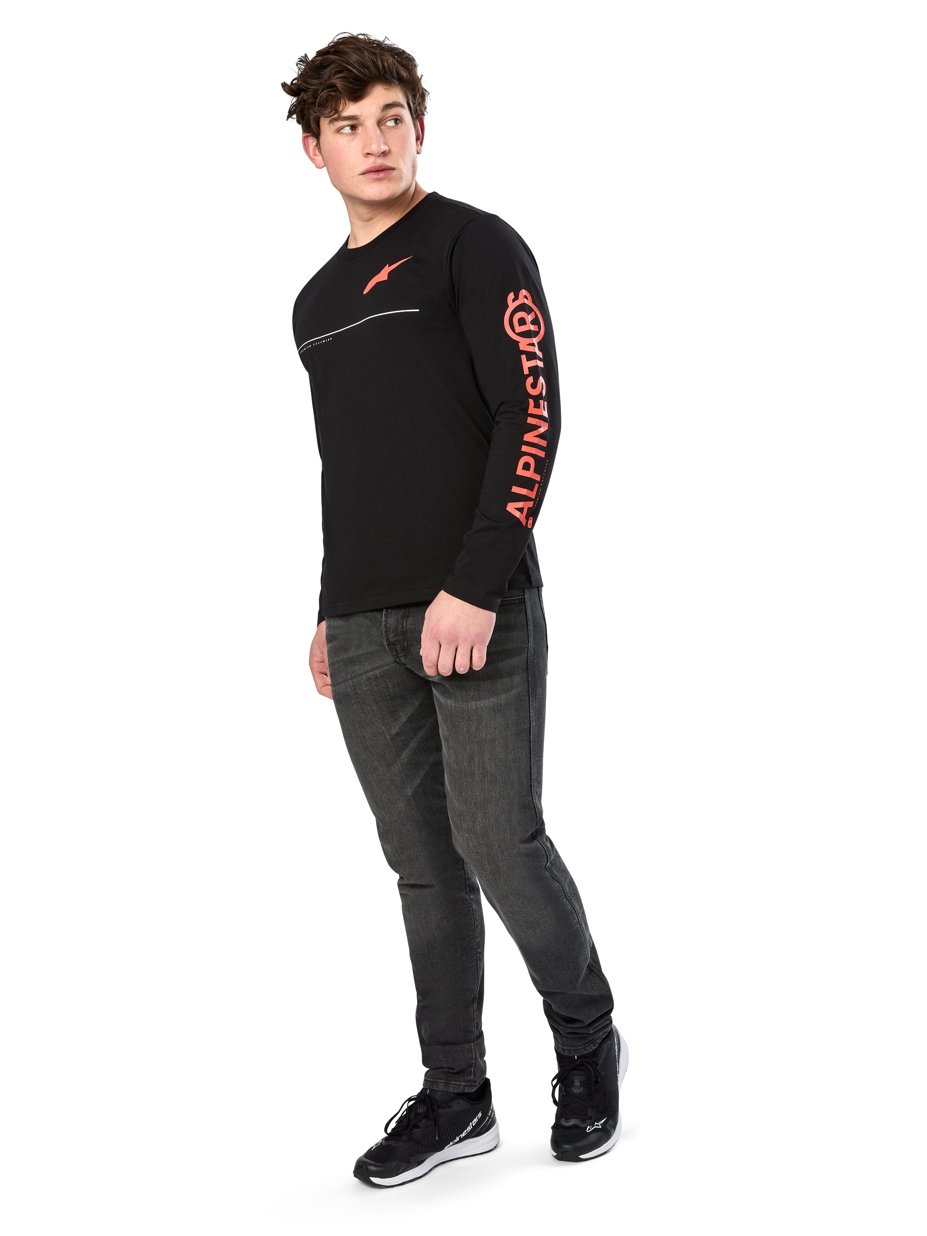Male model wearing Alpinestars Committed CSF Tee, Long Sleeve T-Shirt, Black, orange-red chest logo and sleeve branding, paired with dark gray jeans and black sneakers, casual motorsport-inspired streetwear