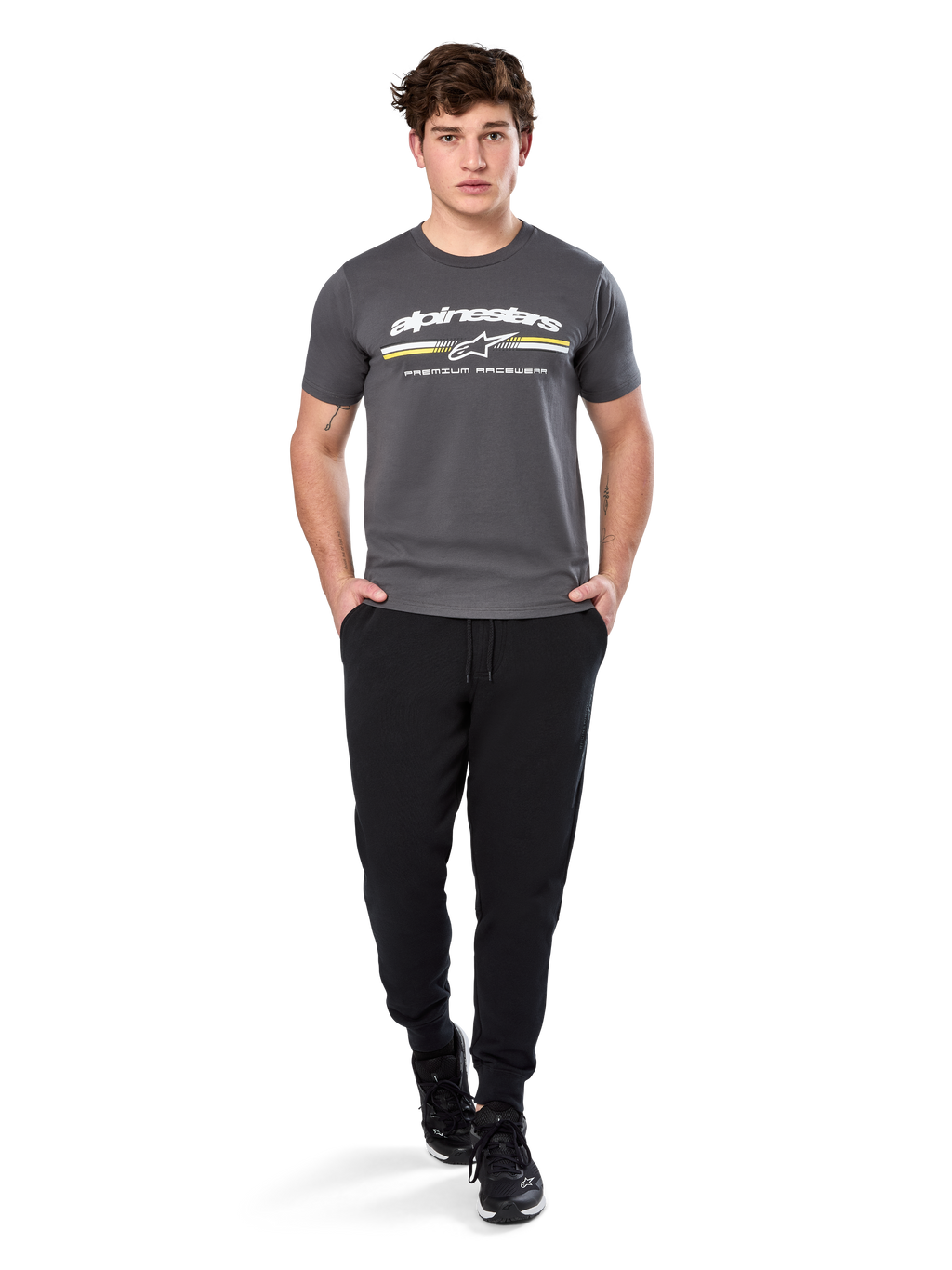 Man wearing Alpinestars Prevail Csf Tee, Short Sleeve T-Shirt, Charcoal, featuring white Alpinestars logo and tri-color stripe with yellow accents, paired with black joggers and sneakers, casual motorsport streetwear
