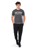Man wearing Alpinestars Prevail Csf Tee, Short Sleeve T-Shirt, Charcoal, featuring white Alpinestars logo and tri-color stripe with yellow accents, paired with black joggers and sneakers, casual motorsport streetwear