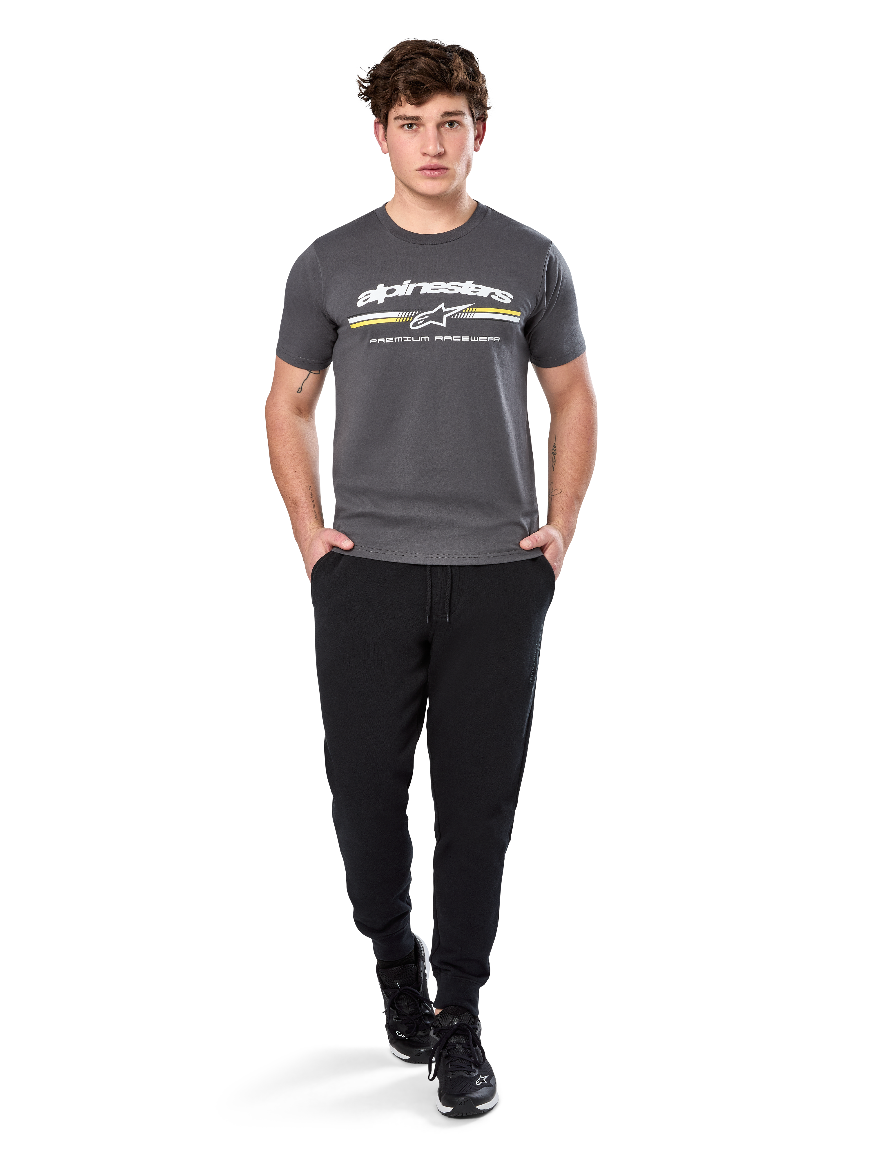 Man wearing Alpinestars Prevail Csf Tee, Short Sleeve T-Shirt, Charcoal, featuring white Alpinestars logo and tri-color stripe with yellow accents, paired with black joggers and sneakers, casual motorsport streetwear