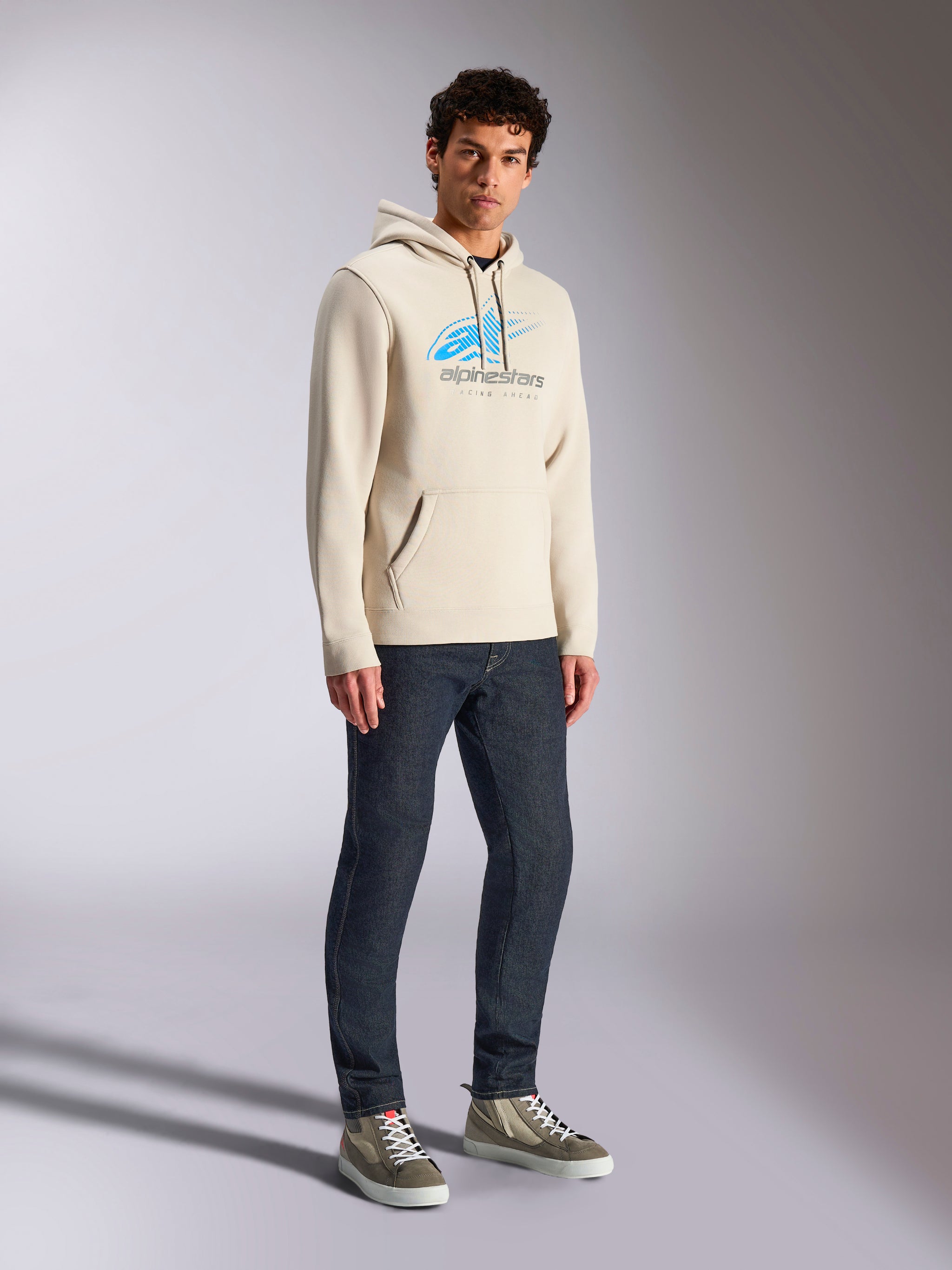 Alpinestars Pulse Hoodie, Off White casual streetwear, hooded pullover fleece featuring a blue and grey Astars logo with speed line fade, kangaroo pocket, worn by model with dark denim jeans and high-top sneakers.