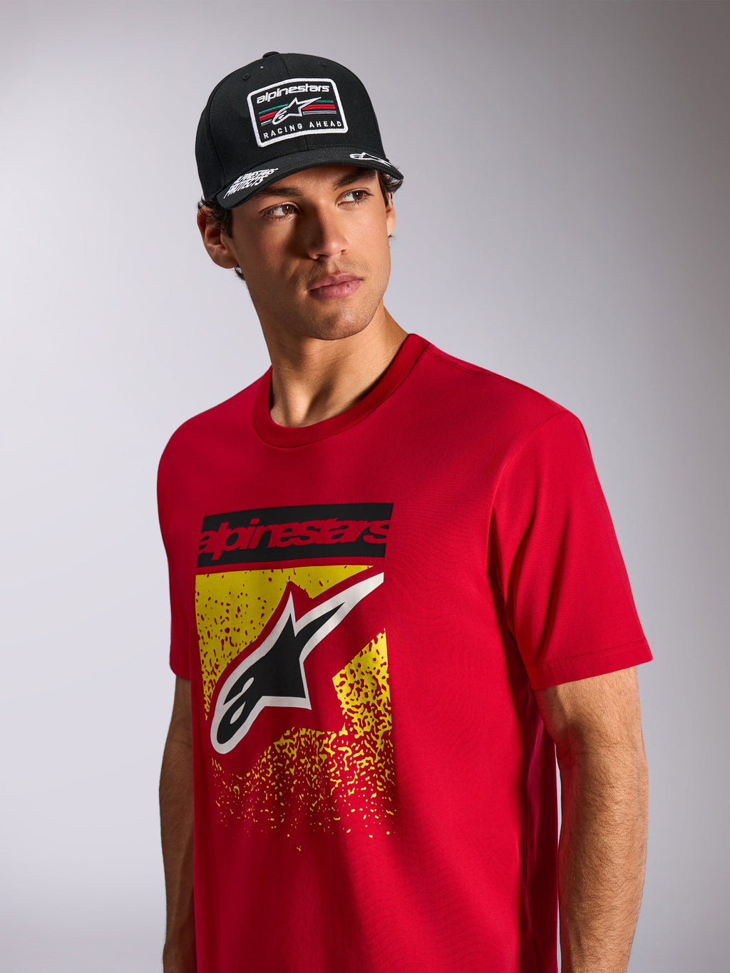Alpinestars Noka CSF Tee, Short Sleeve T-Shirt, Red, featuring a front Astars logo with yellow and red MX gradation, worn with a black Alpinestars 'Racing Ahead' cap, casual motorsport streetwear