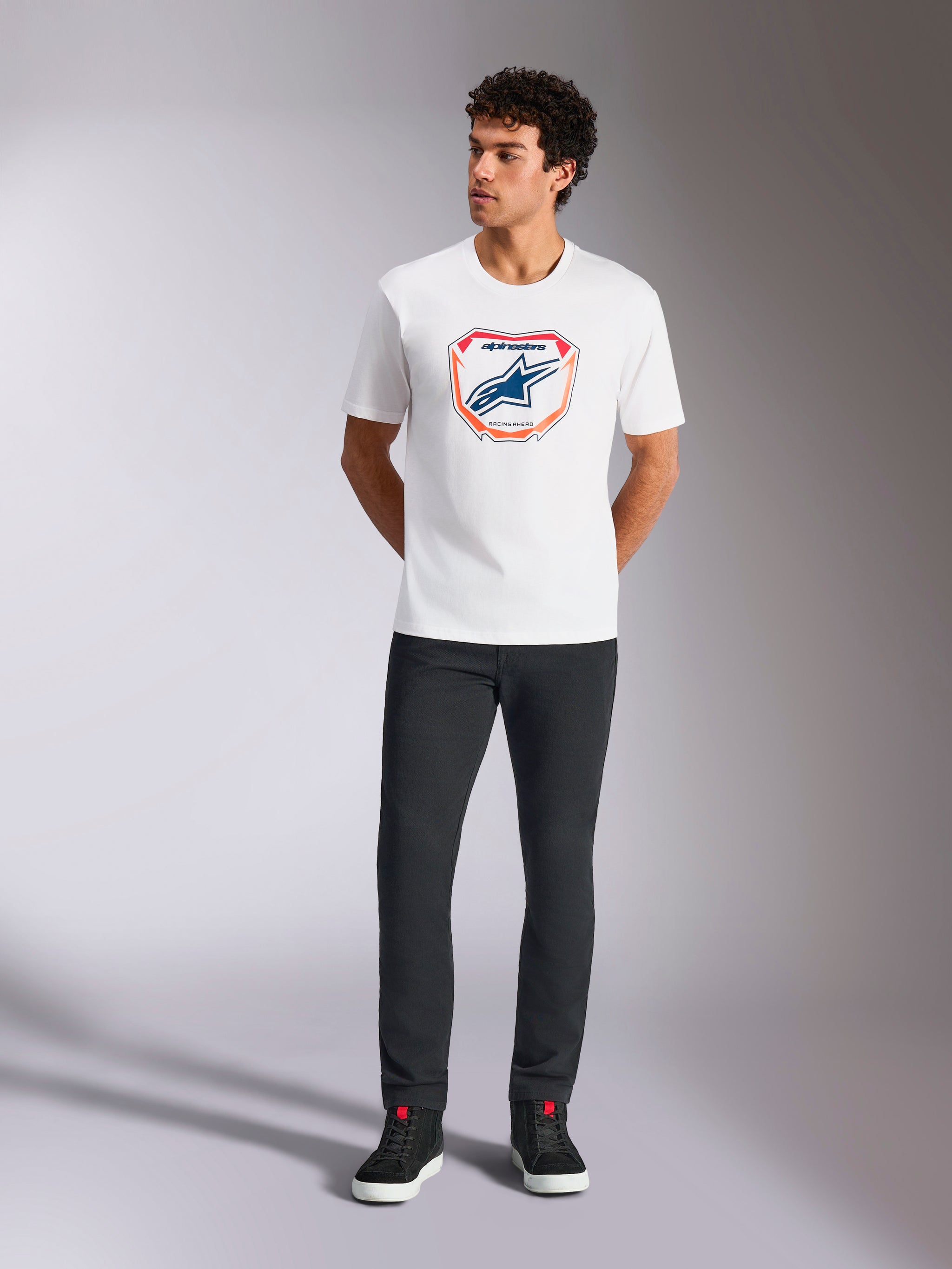 Alpinestars Plate CSF Tee, Short Sleeve T-Shirt, White, featuring a front Astars MX plate graphic with orange and blue accents, worn by a model with black pants and sneakers, Casual Motorsport Streetwear