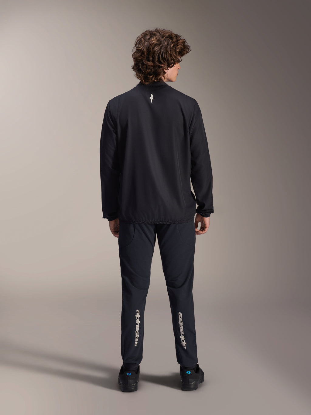 Alpinestars A-Aria Windbreaker Jacket, Black, back view, lightweight wind-resistant cycling jacket with a small white logo on the upper back, worn with matching black pants featuring vertical Alpinestars branding on the calves, performance mountain bike gear