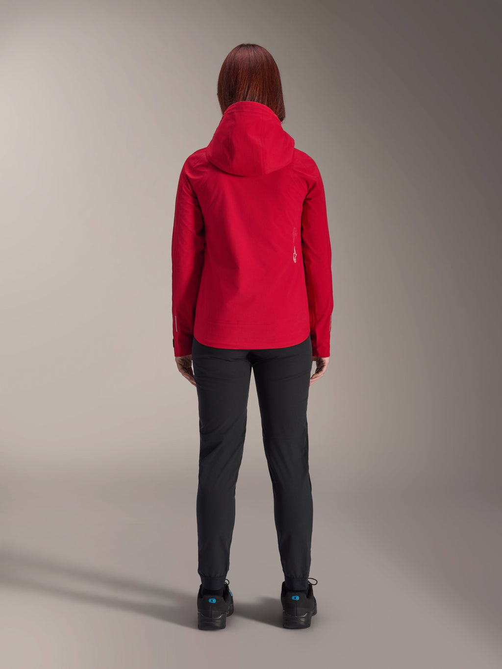 Back view of a woman wearing the Alpinestars Stella A-Dura Elite Waterproof Jacket in Red Fluo, styled with black pants and shoes, featuring a protective hood and dropped tail hem for mountain biking.
