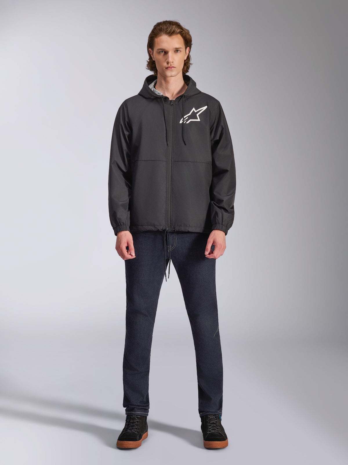 Alpinestars Speeder Chromium Windbreak Jacket, Black with White Astars Logo, worn by model with dark blue jeans and black sneakers, featuring a hooded design and front zipper, casual motorsport-inspired streetwear