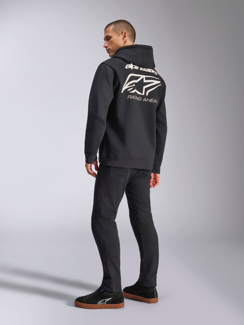 Model wearing Alpinestars Formulation Hoodie, Black, back view featuring large white Alpinestars logo and 'RACING AHEAD' wordmark, casual motorsport-inspired streetwear paired with black pants and black sneakers