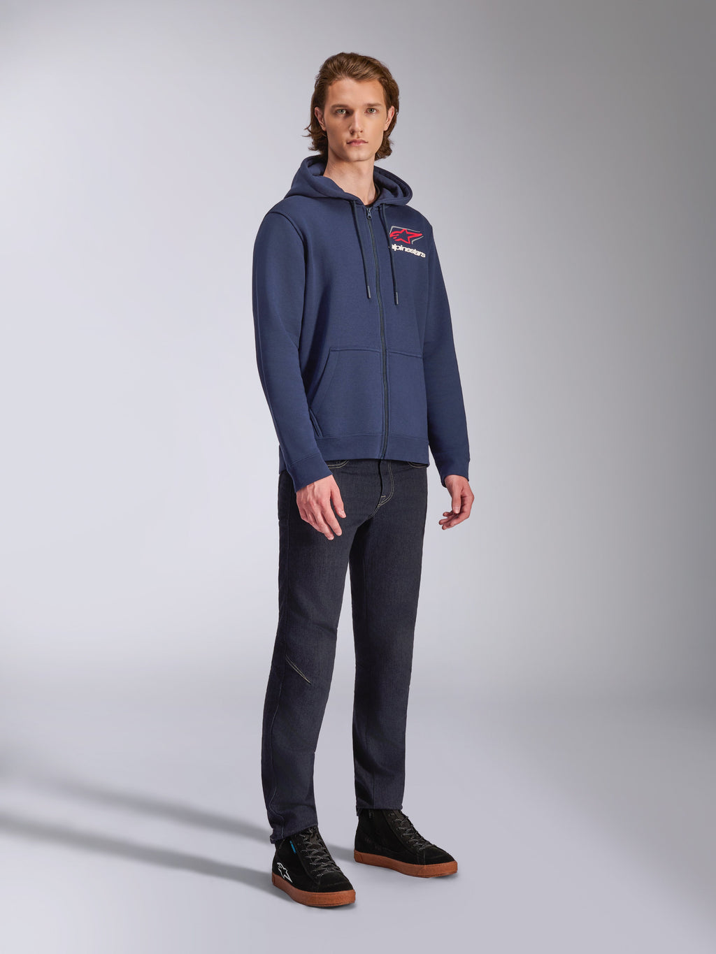 Male model wearing Alpinestars Formulation Hoodie, navy blue zip-front poly fleece, red and white logo on the chest, paired with dark jeans and black sneakers, casual motorsport-inspired streetwear
