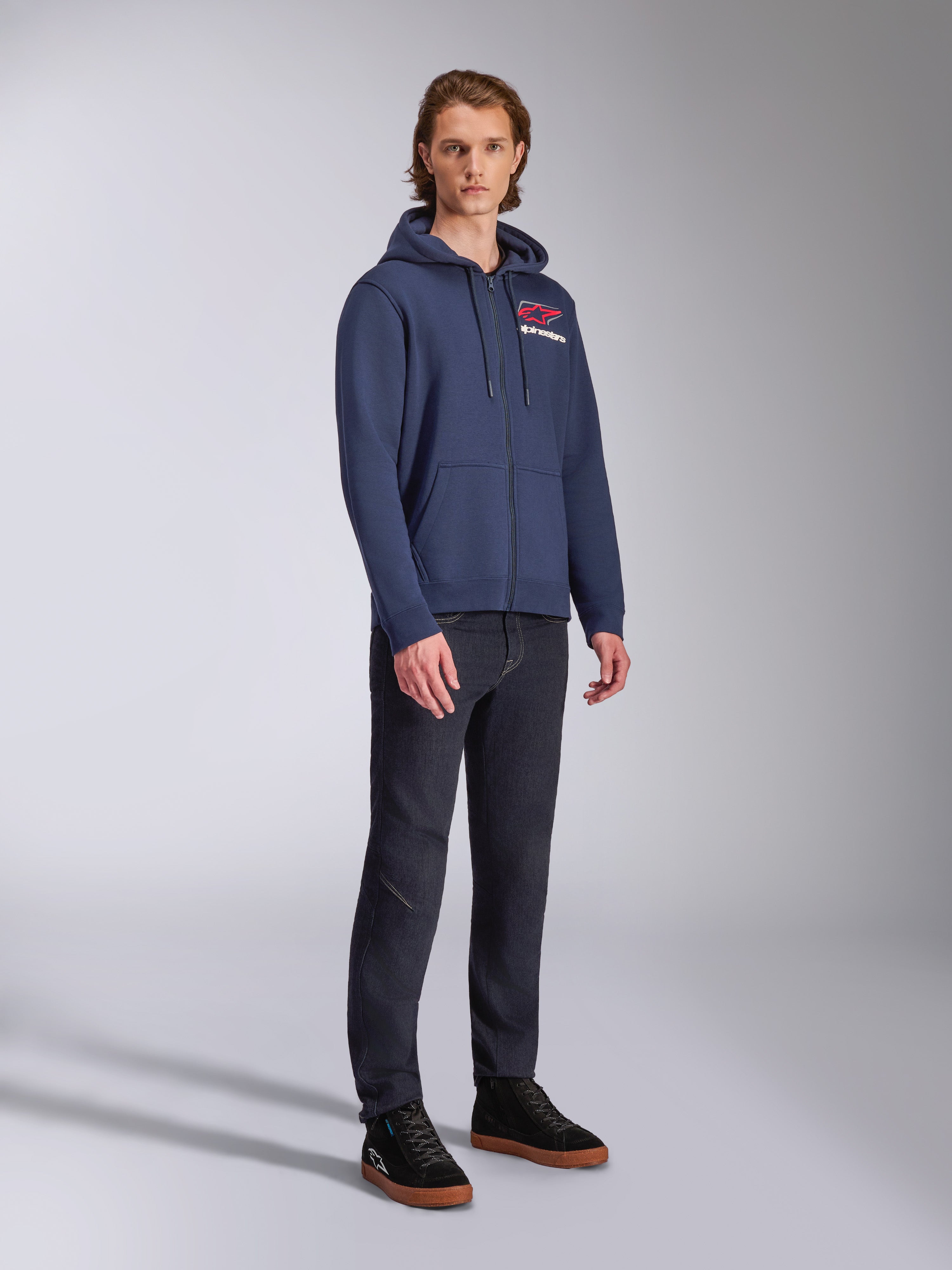 Male model wearing Alpinestars Formulation Hoodie, navy blue zip-front poly fleece, red and white logo on the chest, paired with dark jeans and black sneakers, casual motorsport-inspired streetwear