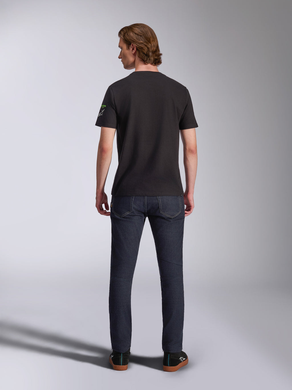 Alpinestars Entitled CSF Tee, Short Sleeve T-Shirt, Black, back view of a male model wearing the shirt with dark blue denim jeans, featuring a small white and green logo on the left sleeve, casual motorsport-inspired streetwear