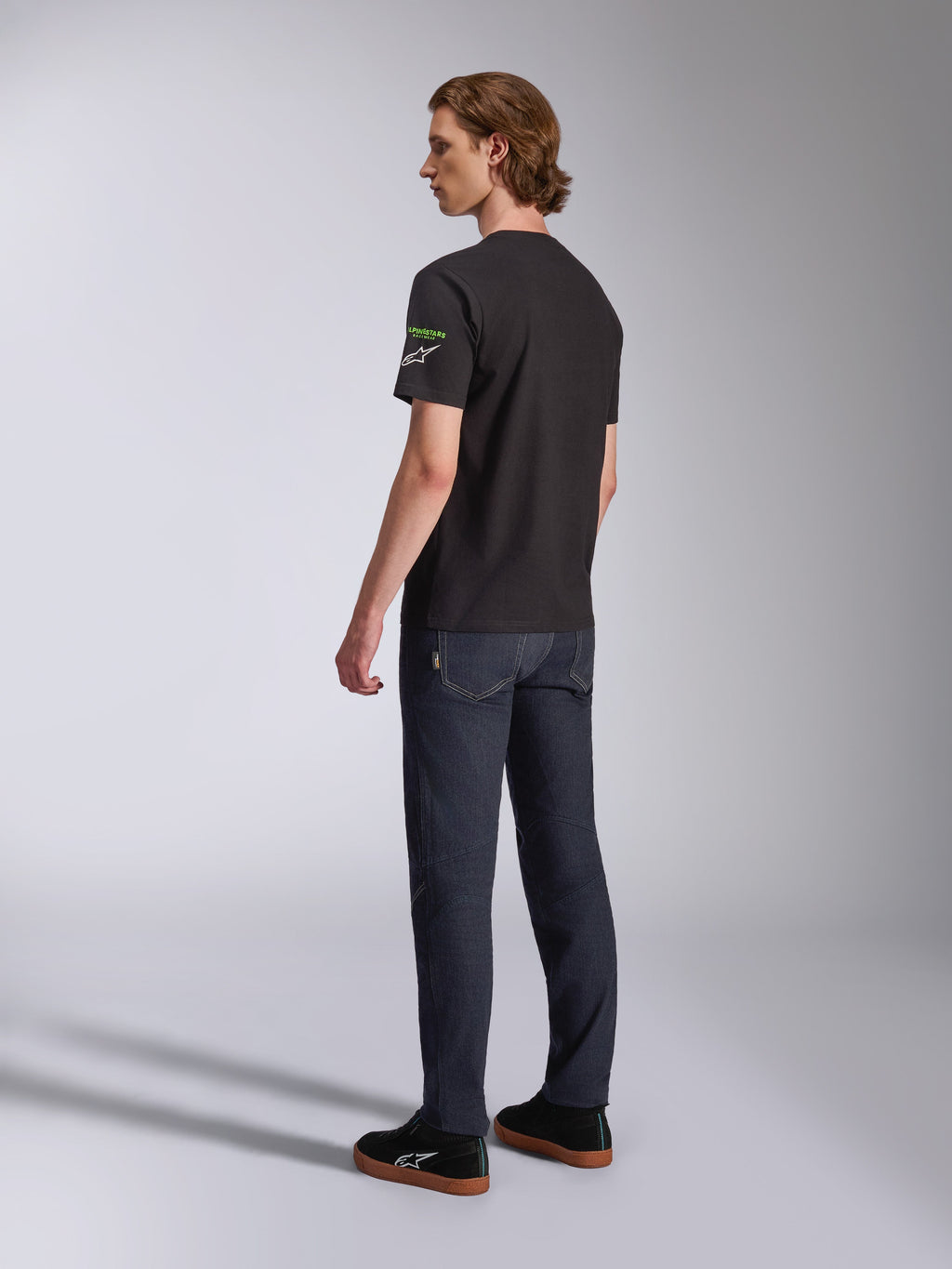Alpinestars Entitled CSF Tee, Short Sleeve T-Shirt, Black, back-side profile of model wearing the tee with green and white sleeve logos, paired with dark denim and black sneakers, casual motorsport streetwear