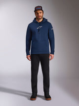 Male model wearing the Alpinestars Frontal Hoodie in Navy, pullover fleece with a white Astars logo on the chest and word logo on the sleeve, featuring a kangaroo pocket and drawstring hood, paired with black pants and a branded cap, casual motorsport streetwear
