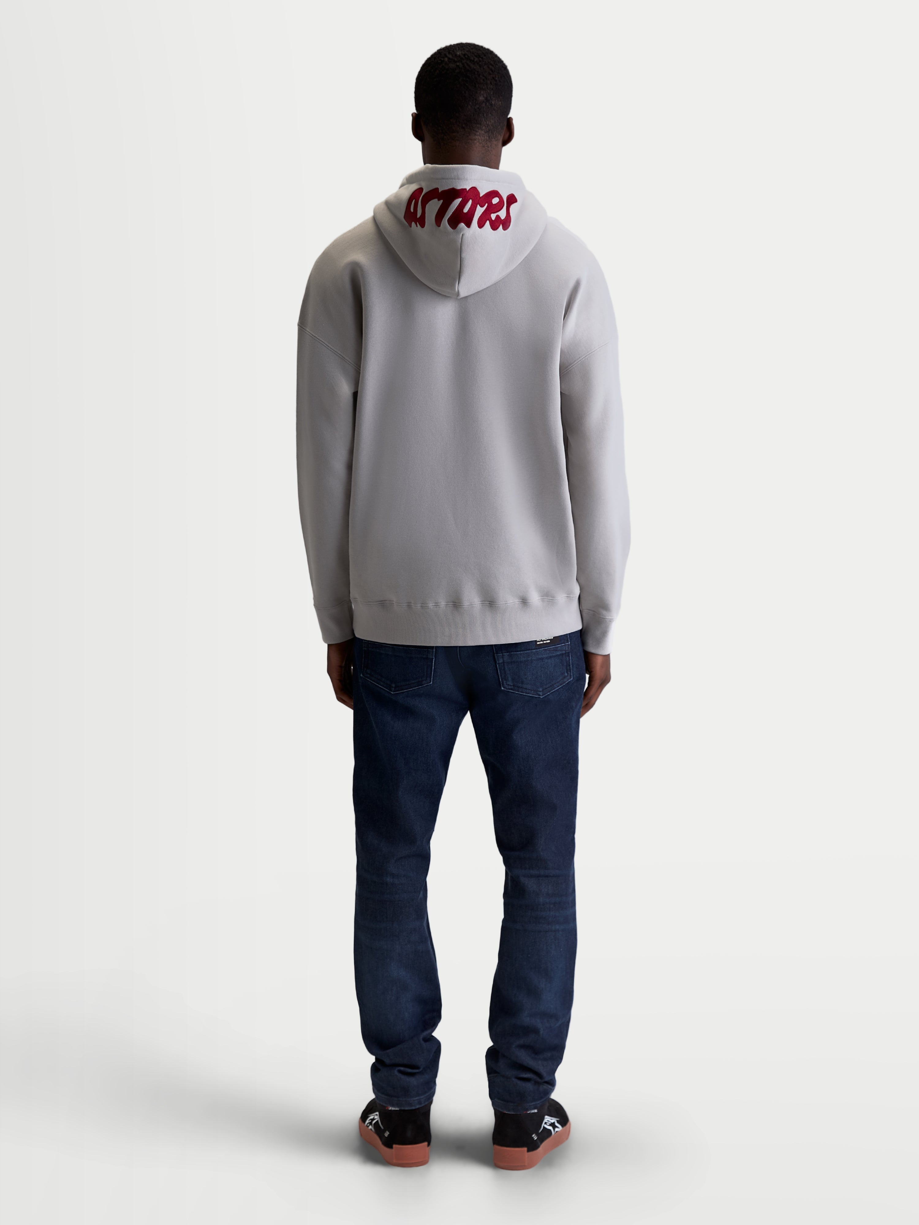 Alpinestars Pioneer Oversized Hoodie, Casual Sportswear Zip Fleece for Men and Women, Cement Gray and Red, Rear view on model highlighting red 'ASTARS' embroidered logo on the hood and relaxed oversized fit.