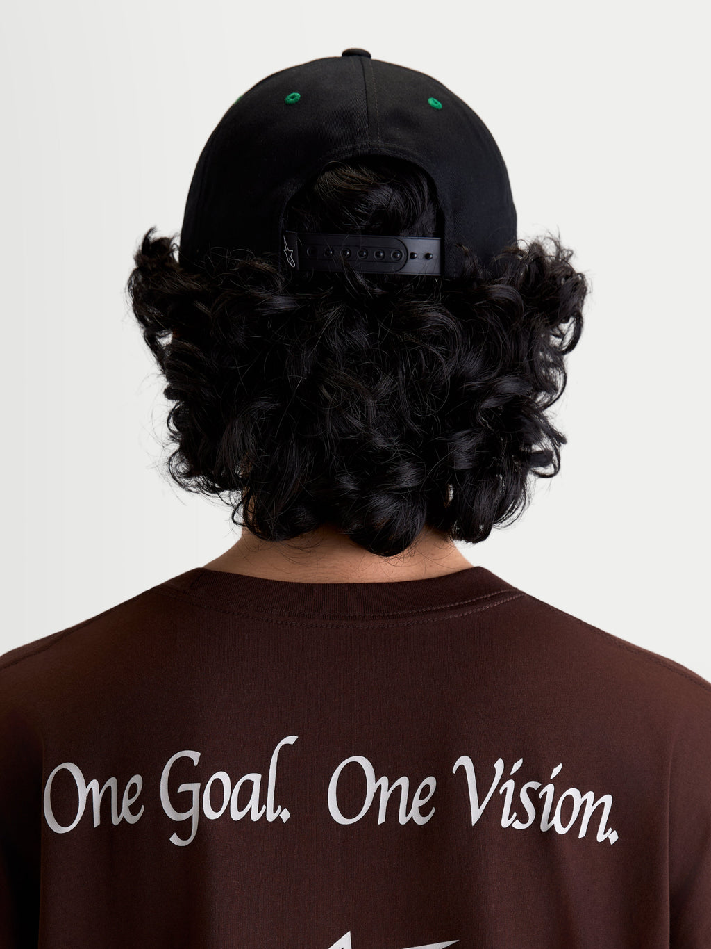 Alpinestars Retro Snapback Hat, Sportswear Snapback Cap, Black and Green, on-model rear view showing adjustable snapback closure and green eyelet details, worn with a brown t-shirt featuring the Alpinestars One Goal. One Vision. brand manifesto.