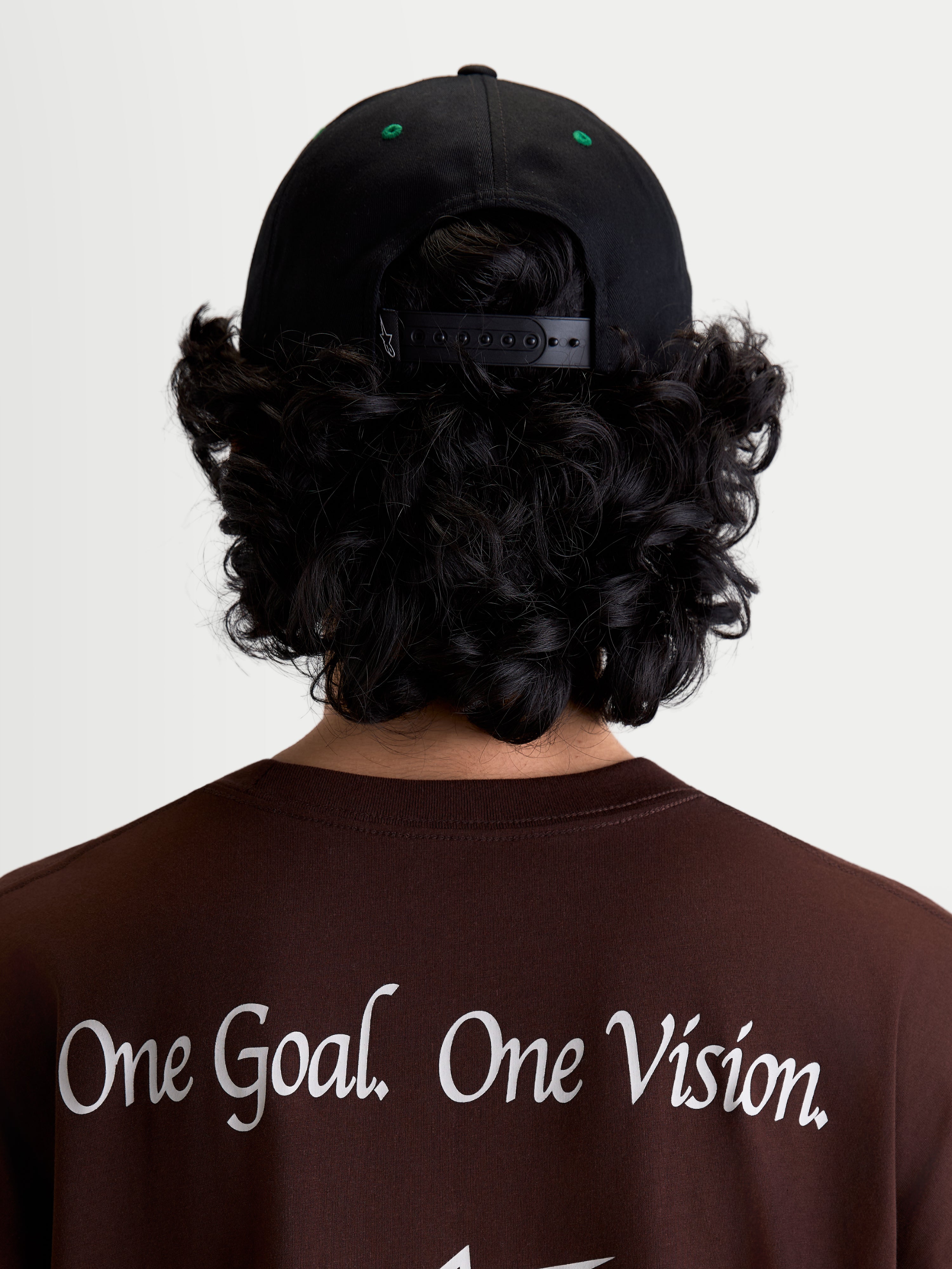 Alpinestars Retro Snapback Hat, Sportswear Snapback Cap, Black and Green, on-model rear view showing adjustable snapback closure and green eyelet details, worn with a brown t-shirt featuring the Alpinestars One Goal. One Vision. brand manifesto.