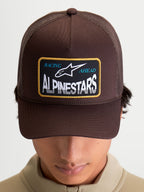Alpinestars Hemmings Trucker Hat, Brown, Sportswear Mesh Cap for Men and Women. Features a structured twill front, breathable poly mesh back, and curved bill. Front design includes a woven Racing Ahead Alpinestars logo patch with white and blue text. On-model front view.