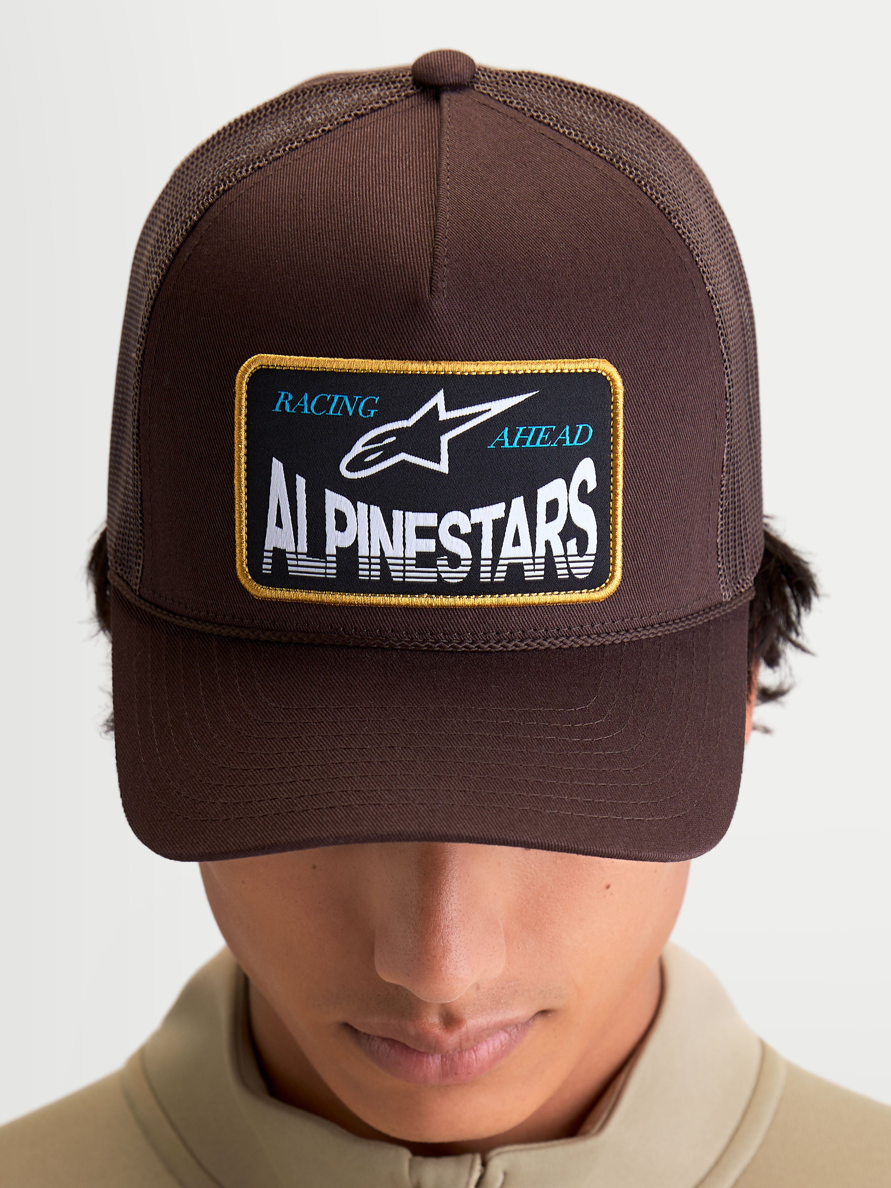 Alpinestars Hemmings Trucker Hat, Brown, Sportswear Mesh Cap for Men and Women. Features a structured twill front, breathable poly mesh back, and curved bill. Front design includes a woven Racing Ahead Alpinestars logo patch with white and blue text. On-model front view.