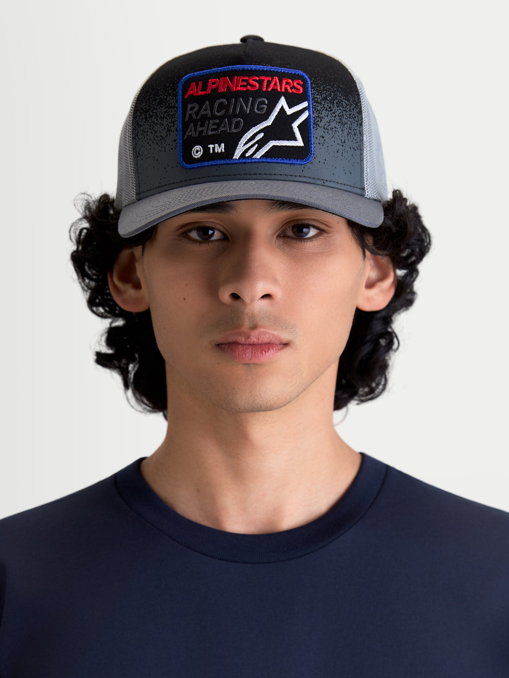 Alpinestars Gravel Trucker Hat, casual sportswear mesh back cap for men and women, Dark Grey and Black, shown on a model. Features a structured crown, curved bill, and a woven front patch with Alpinestars Racing Ahead branding and gravel-texture detailing.