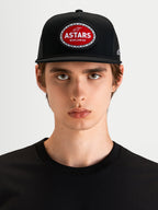 Alpinestars Hotlink Trucker Hat, motorsport-inspired sportswear cap in black, featuring a flat bill, structured crown, and a red and white Astars woven patch with a bike chain border on the front, worn by a model.