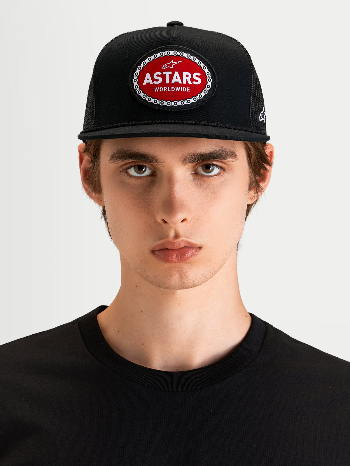 Alpinestars Hotlink Trucker Hat, motorsport-inspired sportswear cap in black, featuring a flat bill, structured crown, and a red and white Astars woven patch with a bike chain border on the front, worn by a model.