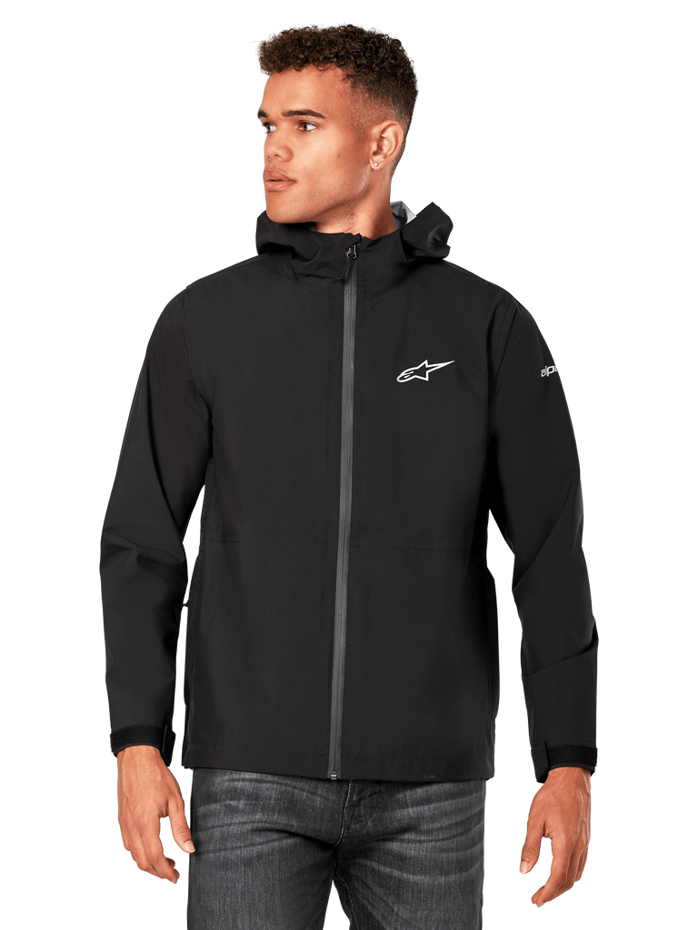 Male model wearing Alpinestars Kascade Rain Jacket, Black, white chest logo and sleeve branding, hooded rain shell with coated front zipper and velcro cuffs, paired with dark jeans, casual motorsport-inspired streetwear