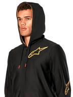 Man wearing Alpinestars Sessions V3 Hoodie, black zip-front fleece with gold star logo on chest, gold mesh-lined hood with tech drawcords, casual motorsport streetwear