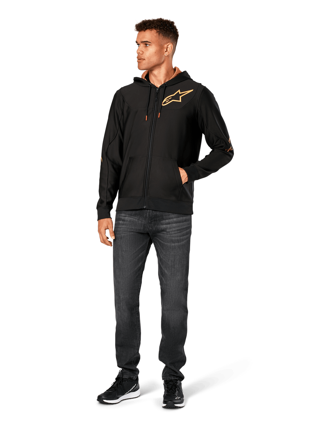 Male model wearing Alpinestars Sessions V3 Hoodie, black zip-front fleece hoodie with gold Astars logo on chest, paired with dark grey jeans and black sneakers, features a gold mesh-lined hood and tech drawcords, casual motorsport streetwear