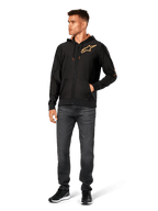Male model wearing Alpinestars Sessions V3 Hoodie, black zip-front fleece hoodie with gold Astars logo on chest, paired with dark grey jeans and black sneakers, features a gold mesh-lined hood and tech drawcords, casual motorsport streetwear