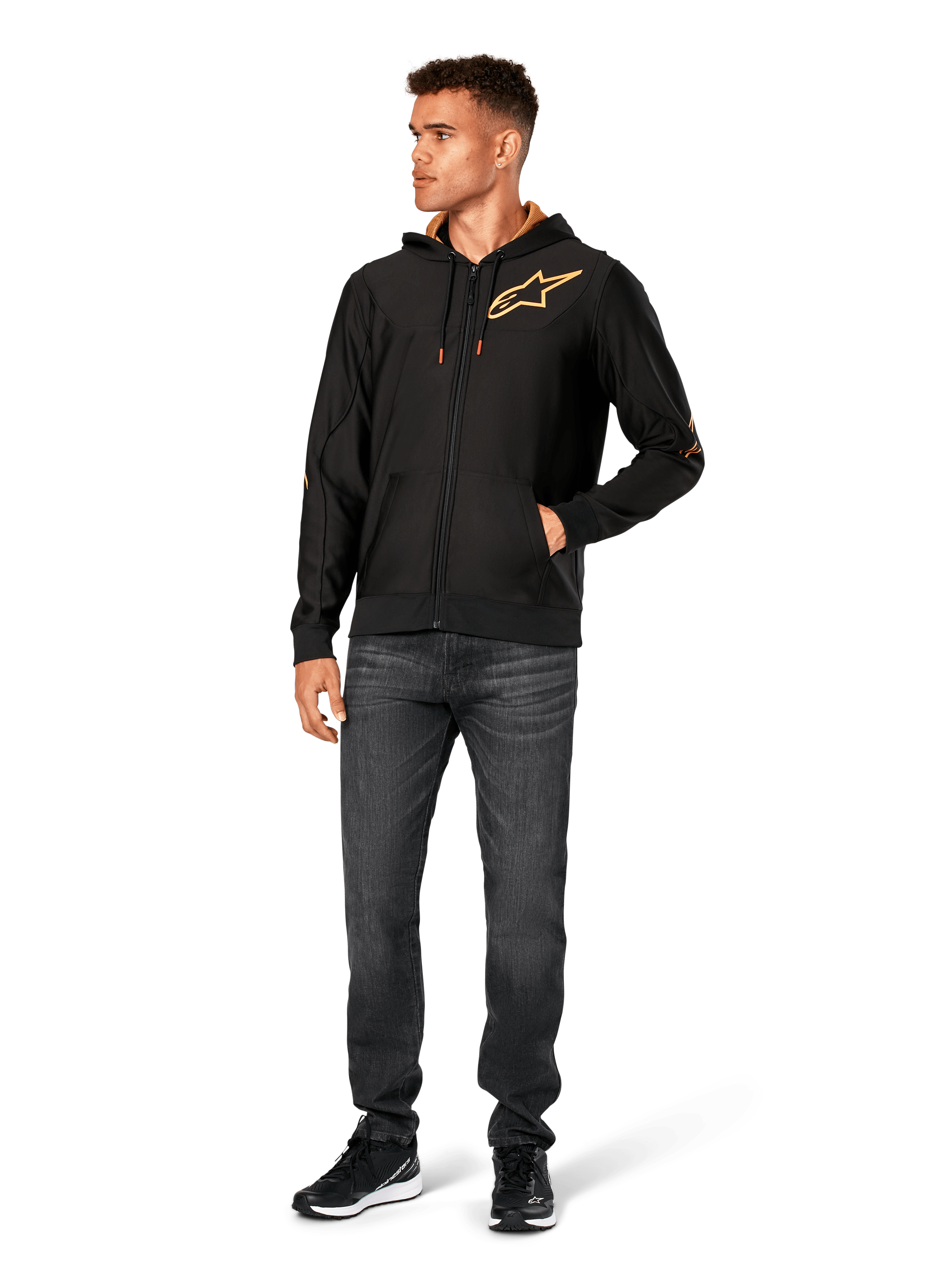 Male model wearing Alpinestars Sessions V3 Hoodie, black zip-front fleece hoodie with gold Astars logo on chest, paired with dark grey jeans and black sneakers, features a gold mesh-lined hood and tech drawcords, casual motorsport streetwear