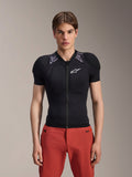Male model wearing Alpinestars A-Motion Plasma Protection Jacket - Short Sleeve, Black with gray camo shoulder padding, white Alpinestars logo on the chest, paired with red pants, slim-fit MTB body armor with a front zipper