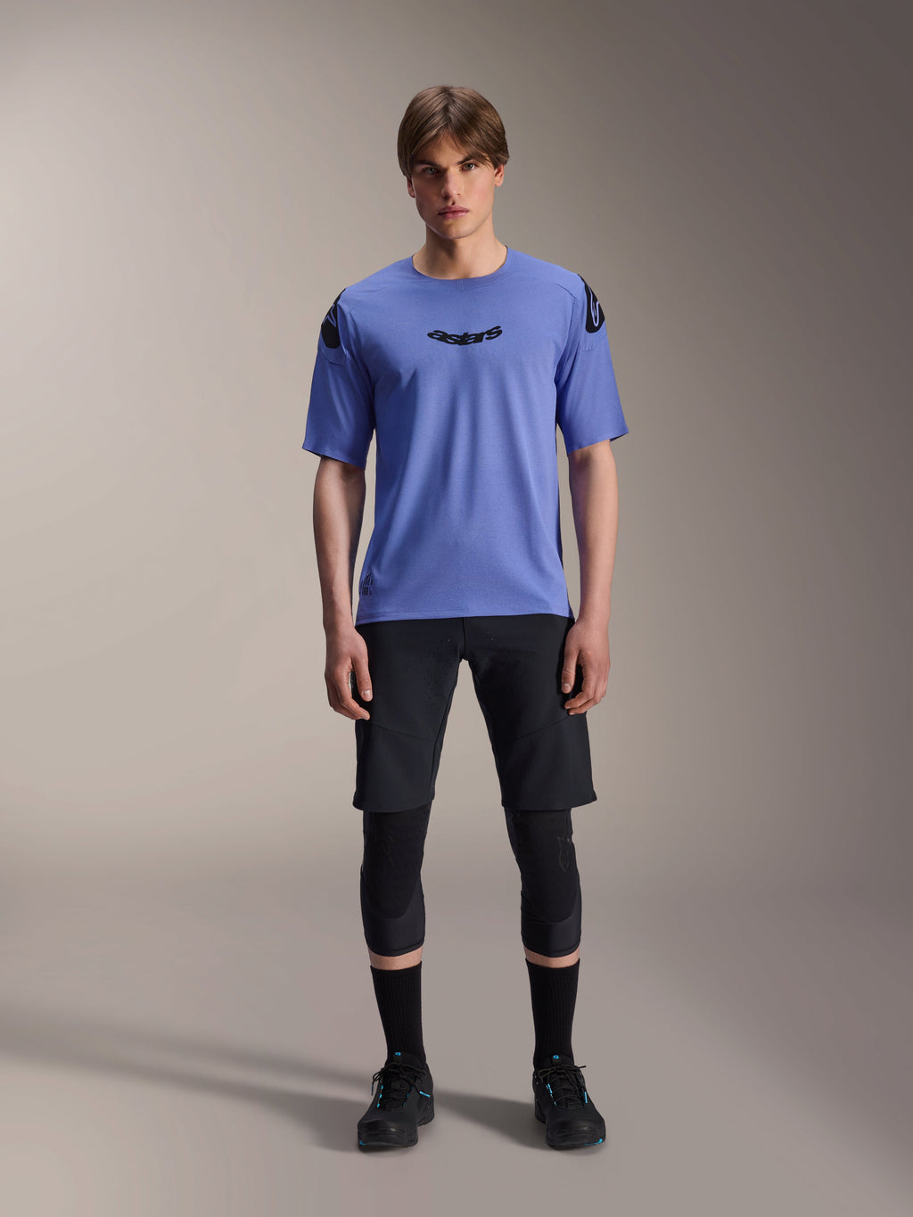 Alpinestars A-Aria Elite Shorts, MTB Mountain Biking Shorts, Black Titanium, worn with blue short-sleeve jersey, knee guards, and black biking shoes, full-body studio shot, performance mountain bike apparel