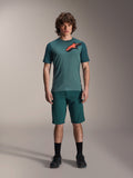 Alpinestars A-Dura Shorts in Dark Pine, men's mountain bike shorts, shown on a model with a matching teal short-sleeve jersey featuring a red and black star logo, black socks and cycling shoes, casual sporty mountain biking apparel