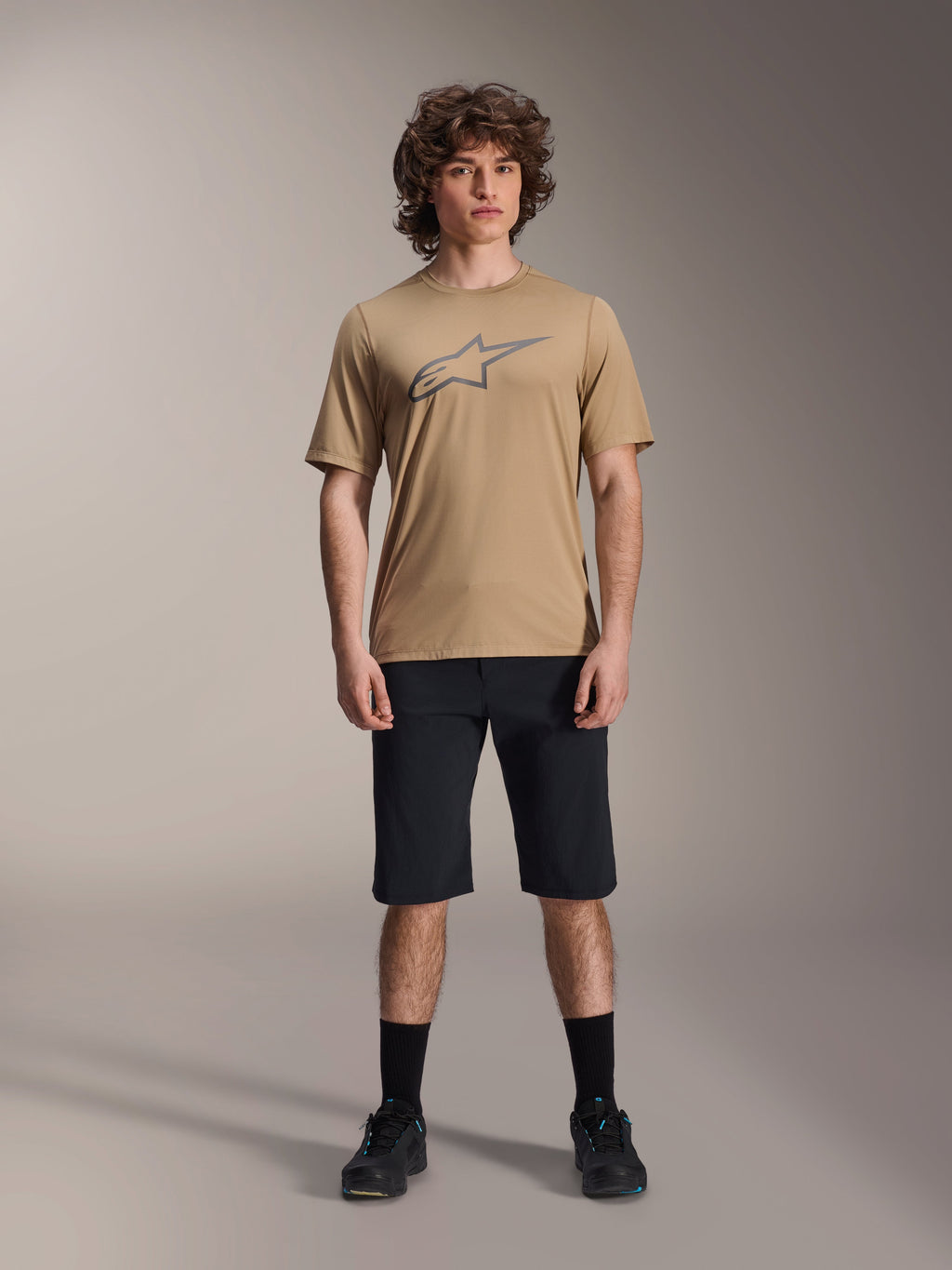 Alpinestars A-Dura Astar Short Sleeve Jersey, Light Brown, worn with black shorts and black shoes, featuring a large outlined Astar logo on the chest, jacquard performance fabric for mountain bike trail riding