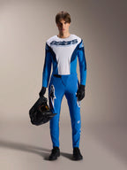 Male model wearing Alpinestars A-Supra Melt MTB kit, white and blue long-sleeve jersey with "astars" wordmark, blue A-Supra Melt pants with white logo, black full-face helmet, black gloves and shoes, coordinated off-road cycling outfit