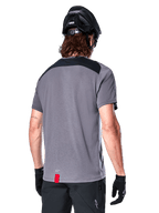 Alpinestars A-Dura Dri Astar Short Sleeve Jersey in Dark Gray, back view showing black mesh panels on the upper back and a longer drop tail, worn with a black MTB helmet and black shorts, technical mountain bike apparel.