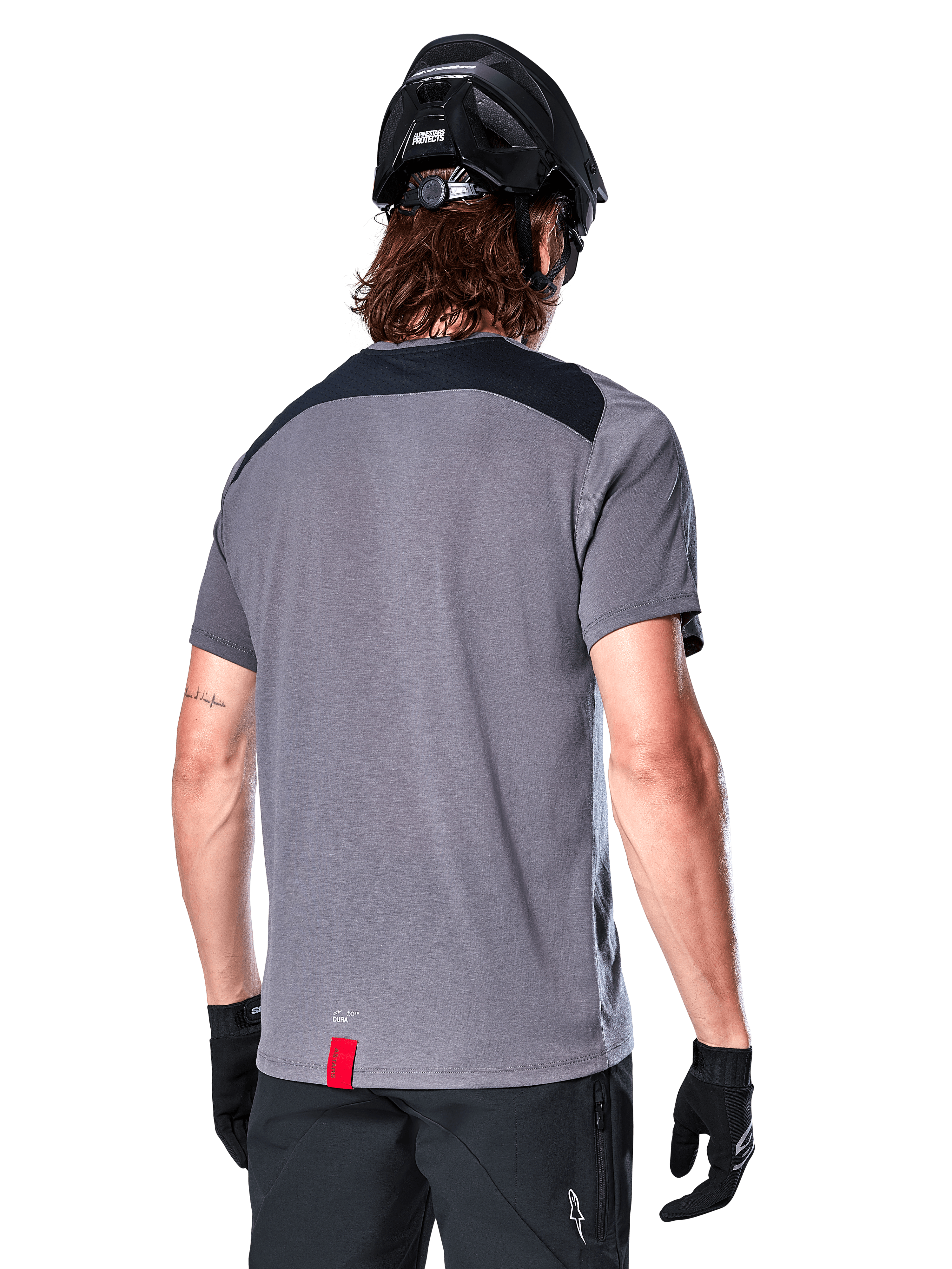 Alpinestars A-Dura Dri Astar Short Sleeve Jersey in Dark Gray, back view showing black mesh panels on the upper back and a longer drop tail, worn with a black MTB helmet and black shorts, technical mountain bike apparel.