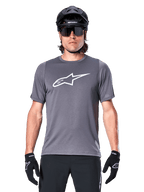 Male mountain biker wearing Alpinestars A-Dura Dri Astar Short Sleeve Jersey, Dark Gray with white Astar logo, paired with a black MTB helmet, sunglasses, and gloves, featuring breathable mesh sleeve panels.