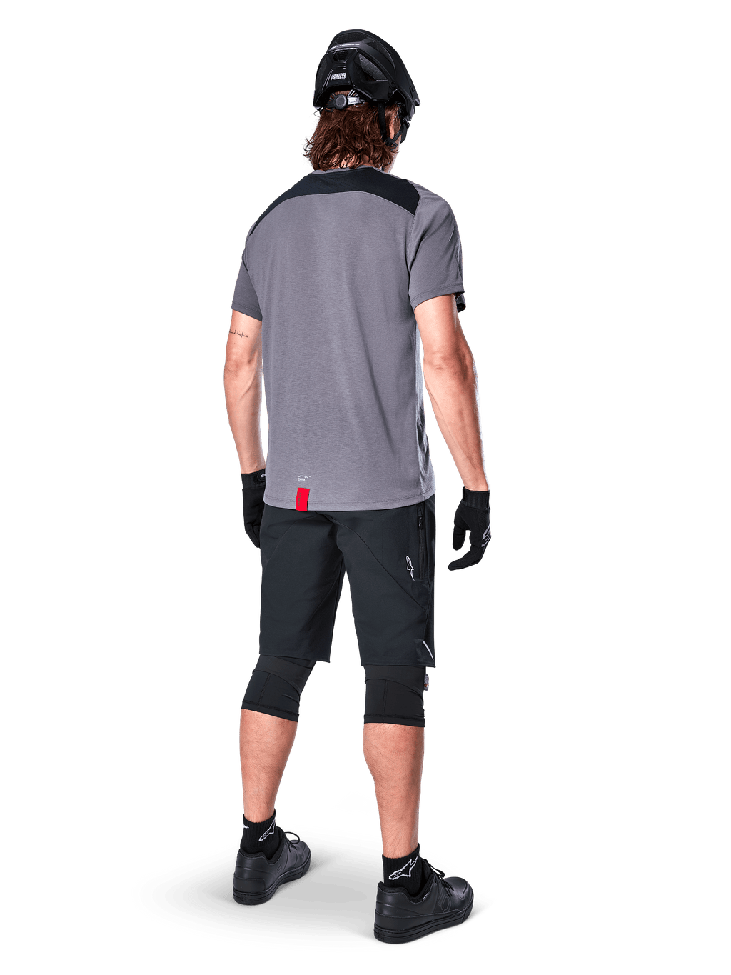 Mountain bike rider seen from behind wearing Alpinestars A-Dura Dri Astar Jersey, Short Sleeve, Dark Gray with black shoulder panels, paired with black MTB shorts, knee pads, and helmet, technical off-road cycling apparel