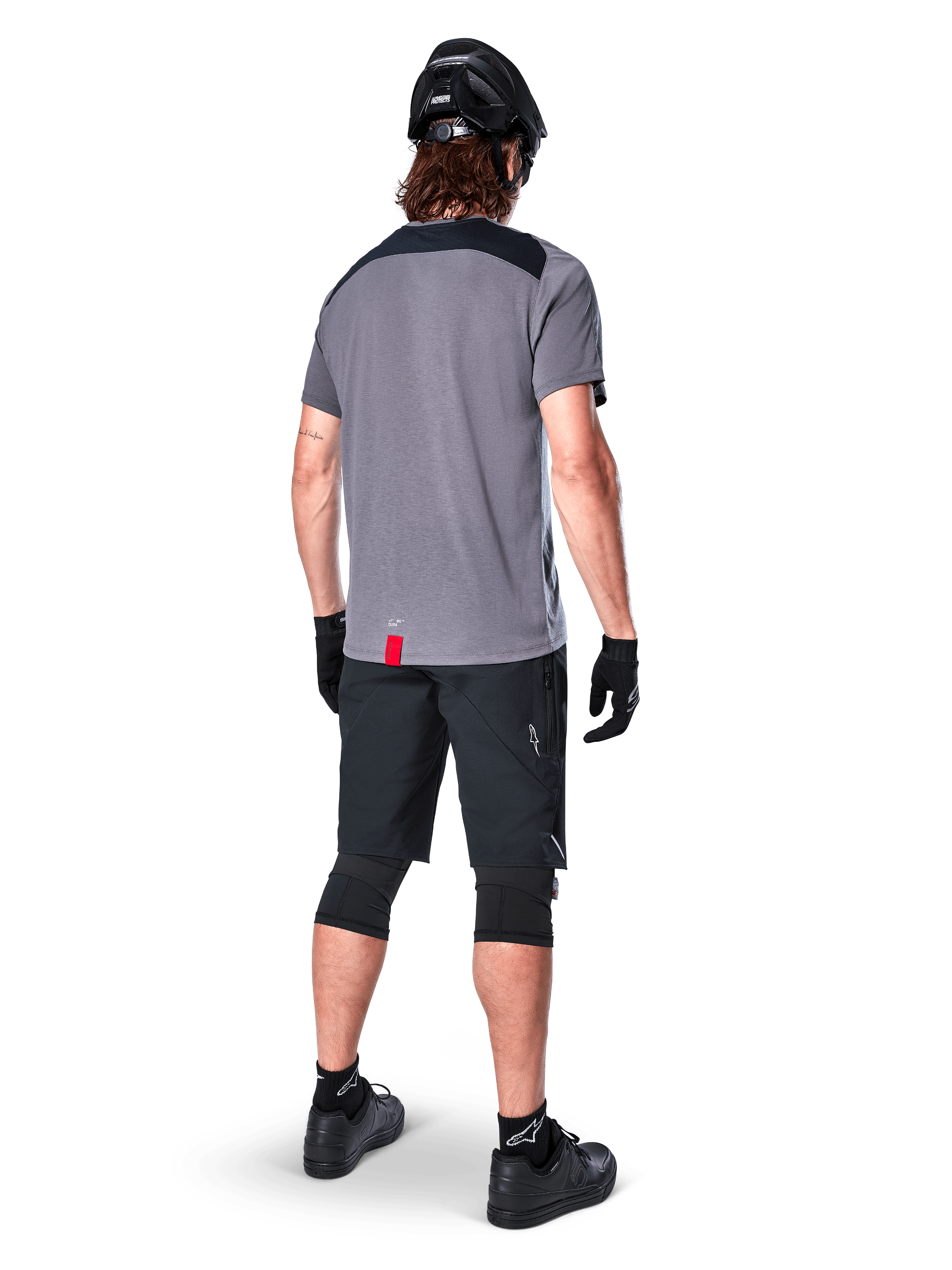 Mountain bike rider seen from behind wearing Alpinestars A-Dura Dri Astar Jersey, Short Sleeve, Dark Gray with black shoulder panels, paired with black MTB shorts, knee pads, and helmet, technical off-road cycling apparel