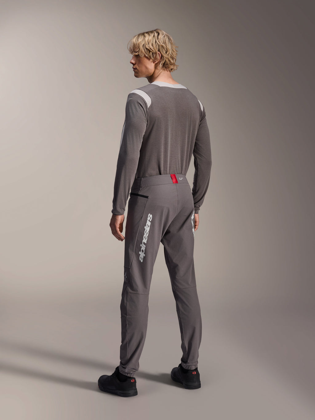 Model from a rear view wearing Alpinestars MTB gear, gray A-Dura Dri Wool Long Sleeve Jersey, Dark Gray A-Dura Elite Pants with white vertical branding and red waistband detail, coordinated cycling outfit for mountain biking