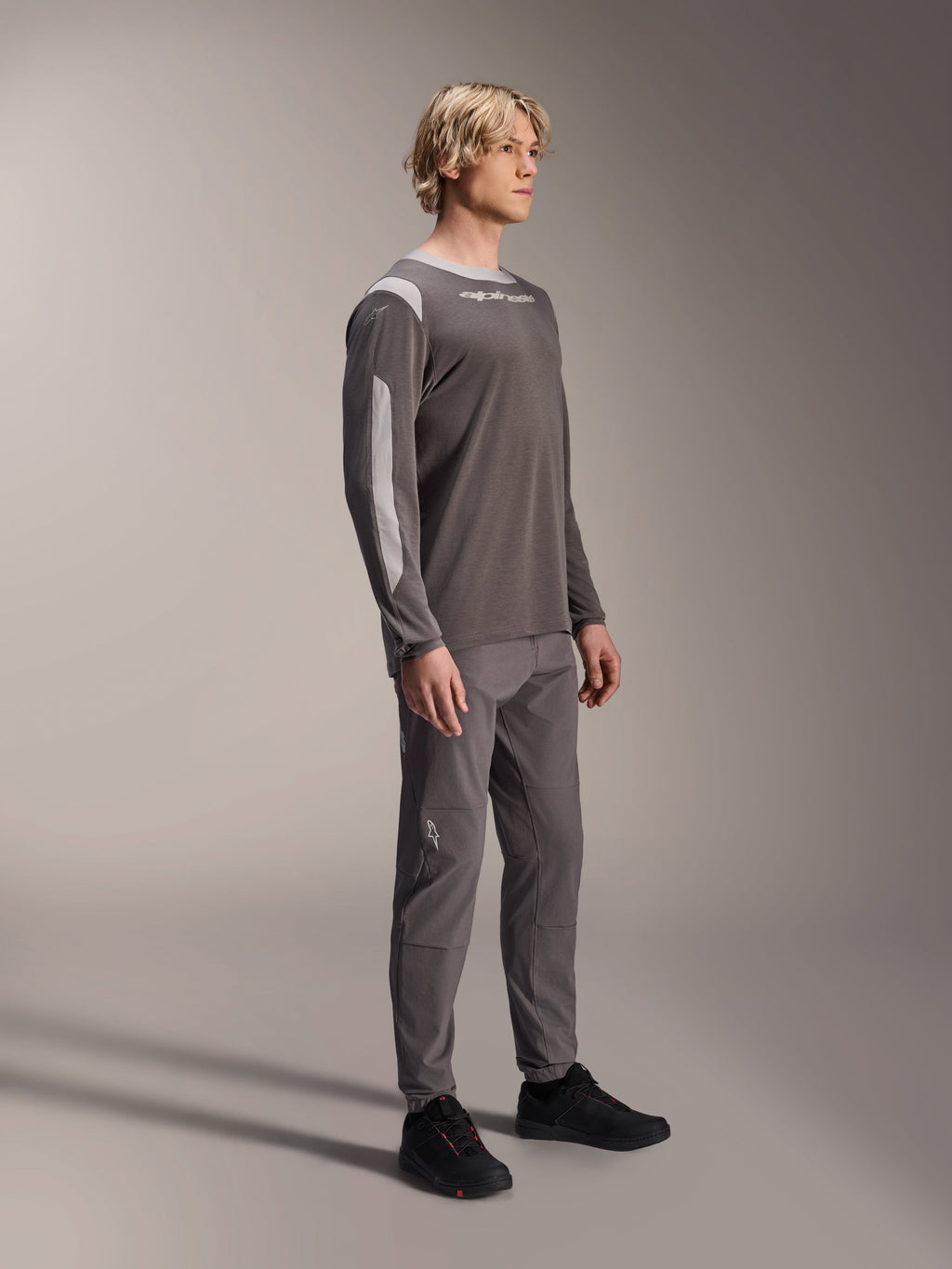 Male model wearing Alpinestars A-Dura Dri Wool Long Sleeve Jersey in Dark Gray, paired with matching gray pants and black shoes, featuring light gray mesh panels on the shoulders and sleeves, technical MTB gear for off-road cycling