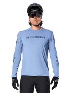 Alpinestars A-Dura Switch Long Sleeve Jersey, Infinity Blue, worn by a man with a black MTB helmet and sunglasses, featuring chest wordmark and sleeve graphics, performance mountain bike apparel