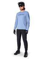 Alpinestars A-Dura Switch Long Sleeve Jersey, Infinity Blue, worn by a rider with black MTB pants, black helmet, and gloves, featuring a chest wordmark logo and star graphics on the sleeves, lightweight cycling gear for trail riding