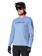 Alpinestars A-Dura Switch Long Sleeve Jersey, Infinity Blue, featuring dark Alpinestars wordmark on the chest and star graphics on the sleeves, worn by a rider with a black mountain bike helmet and sunglasses